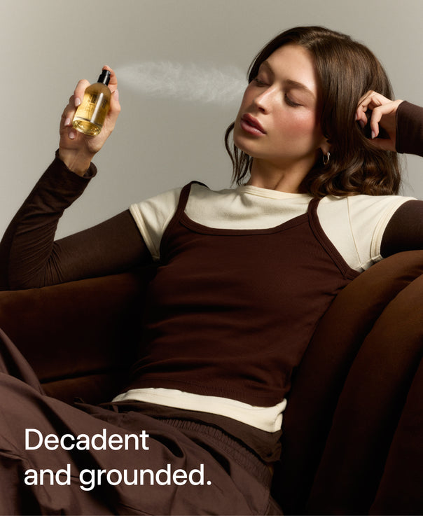 A woman reclining on a brown couch sprays a bottle of fragrance. She wears a brown long-sleeve top over a cream crewneck shirt.